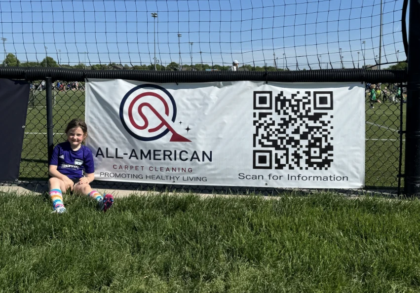 A young KC Stix player beside the All American Carpet Cleaning sponsorship banner at the ballpark